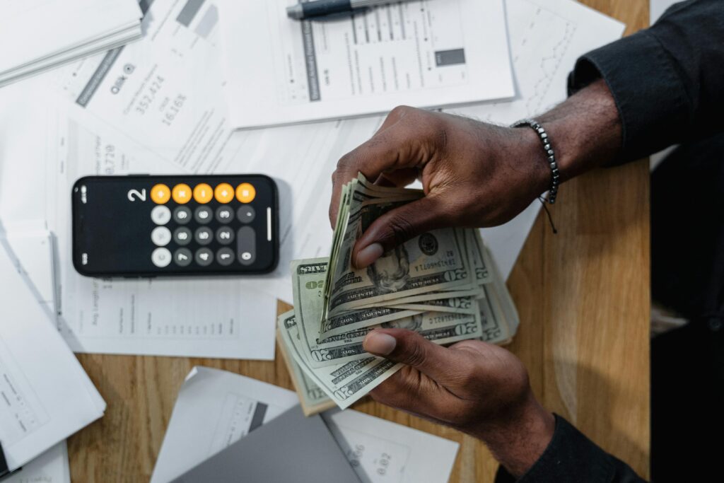 Person counting dollar bills over documents with a smartphone calculator on the desk.